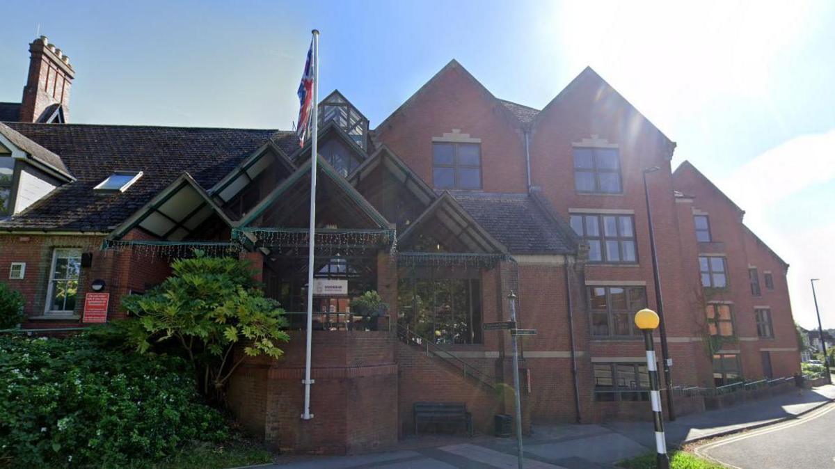 The Wokingham Borough Council building. A Union Jack flies on a flag post outside. It is a large red brick building with gable roofs, and steps lead up to the entrance from the pavement.