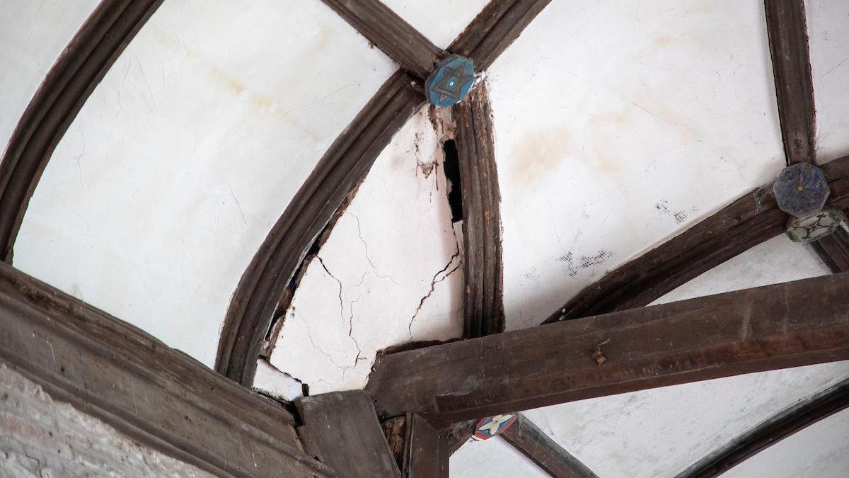 A picture of a roof - it is white plaster with wooden beams.  There are large cracks and holes in one part of the plaster. 