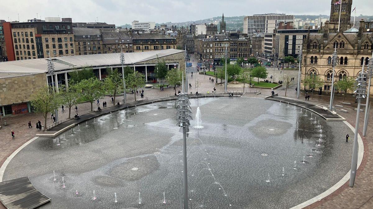 A large public square featuring a circular water feature, with multiple jets spraying water at varying heights. The water gathers to create a mirror effect. Tall light poles surround the fountain, with trees and benches lining the perimeter. People can be seen walking and sitting around the edges of the fountain. In the background, a wider city centre can be seen with a mix of modern and older buildings.