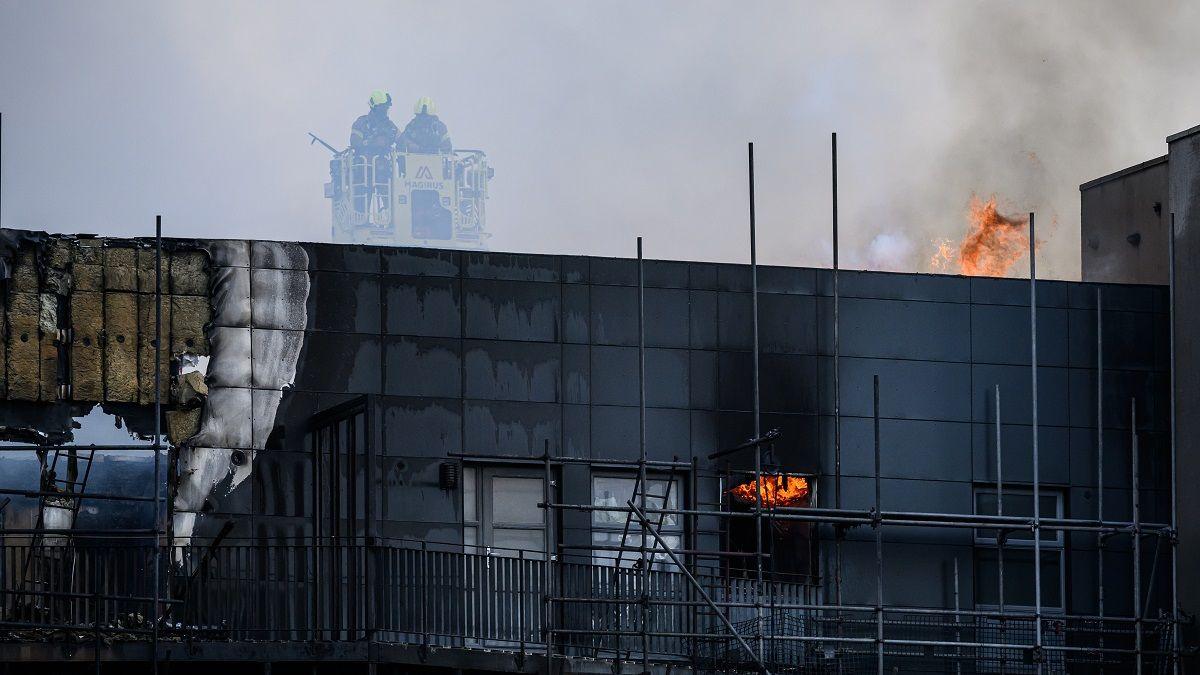 Firefighters on top of the burned building, with flames still coming from the roof and window