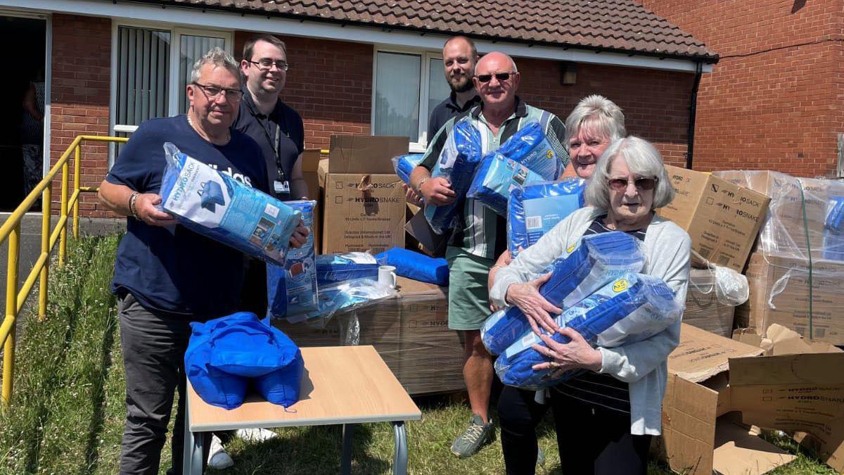 Half a dozen adults stand in two lines surrounded by unpacked boxes. They are holding blue inflatable aqua packs.