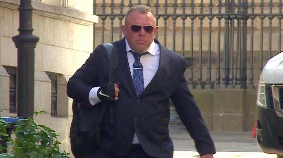 Builder Mark Killick arriving outside Bristol Crown Court. He is wearing a white collared shirt, blue paisley tie with a silver tie pin, a dark suit and sunglasses. He is carrying a black rucksack on one shoulder and walking towards the camera with a straight face. 
