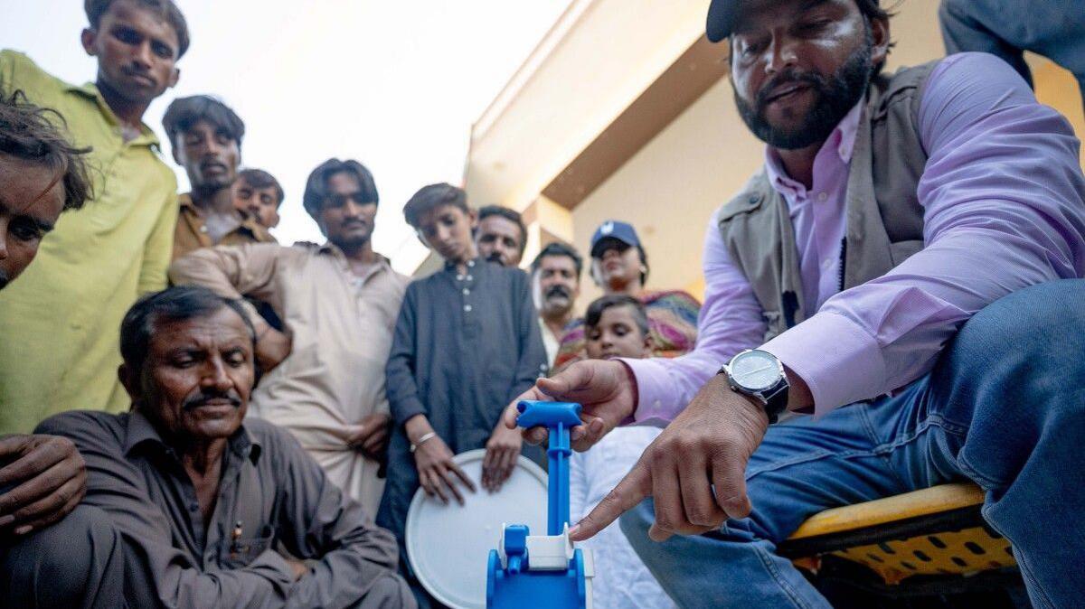 A man is sat down operating a water pump. Behind him a group of people are watching. 