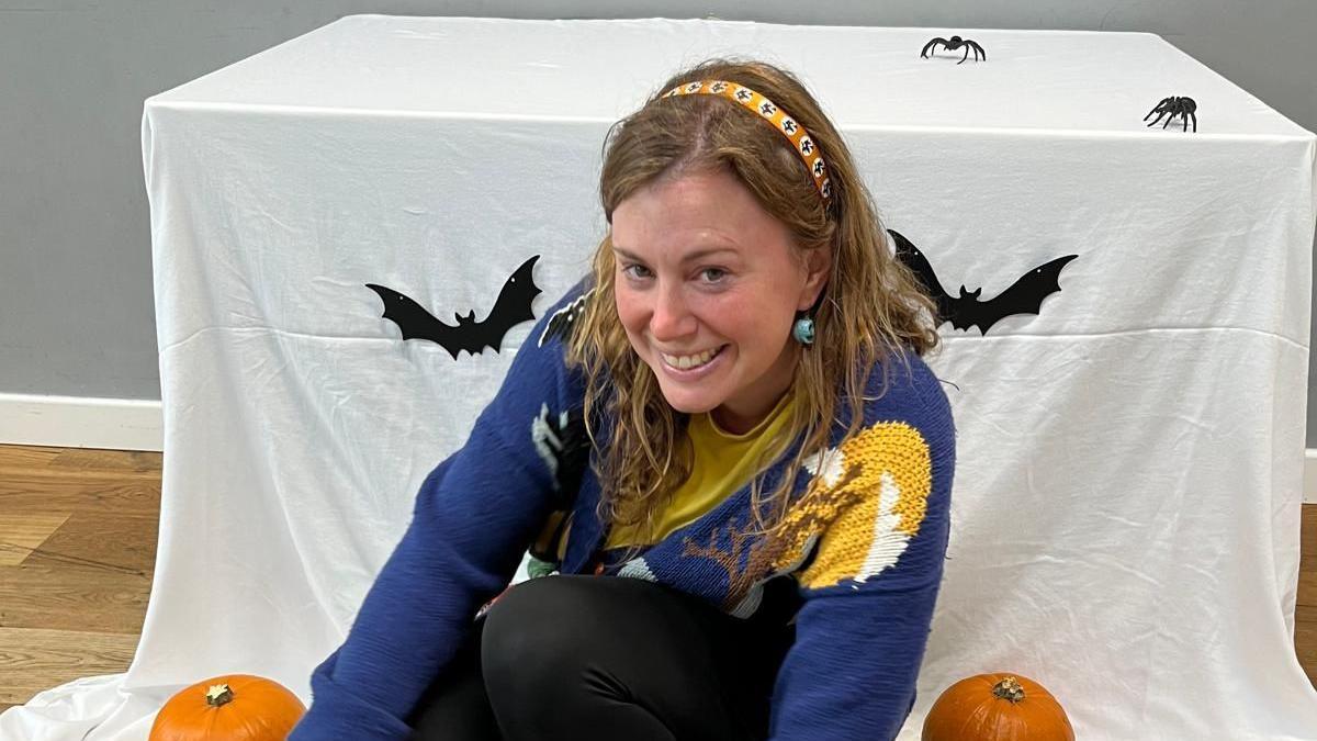 Maia Banks wearing blue cardigan and yellow t-shirt with black trousers. She is sitting in front of a table which has a white sheet over it with Halloween decorations including fake spiders and bats, and pumpkins