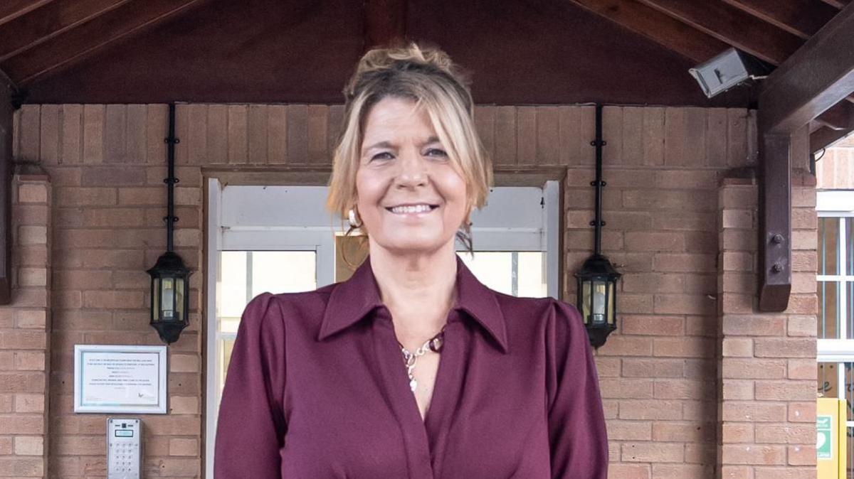 A woman with blonde hair and a maroon blouse standing in front of the entrance to a brick building