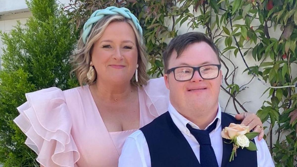 Clare and Joel at a wedding. Joel is wearing a white shirt with a navy waistcoat and tie, dark rimmed glasses and a cream flower. Clare is wearing a blue headband, gold drop earrings and a pink dress. 