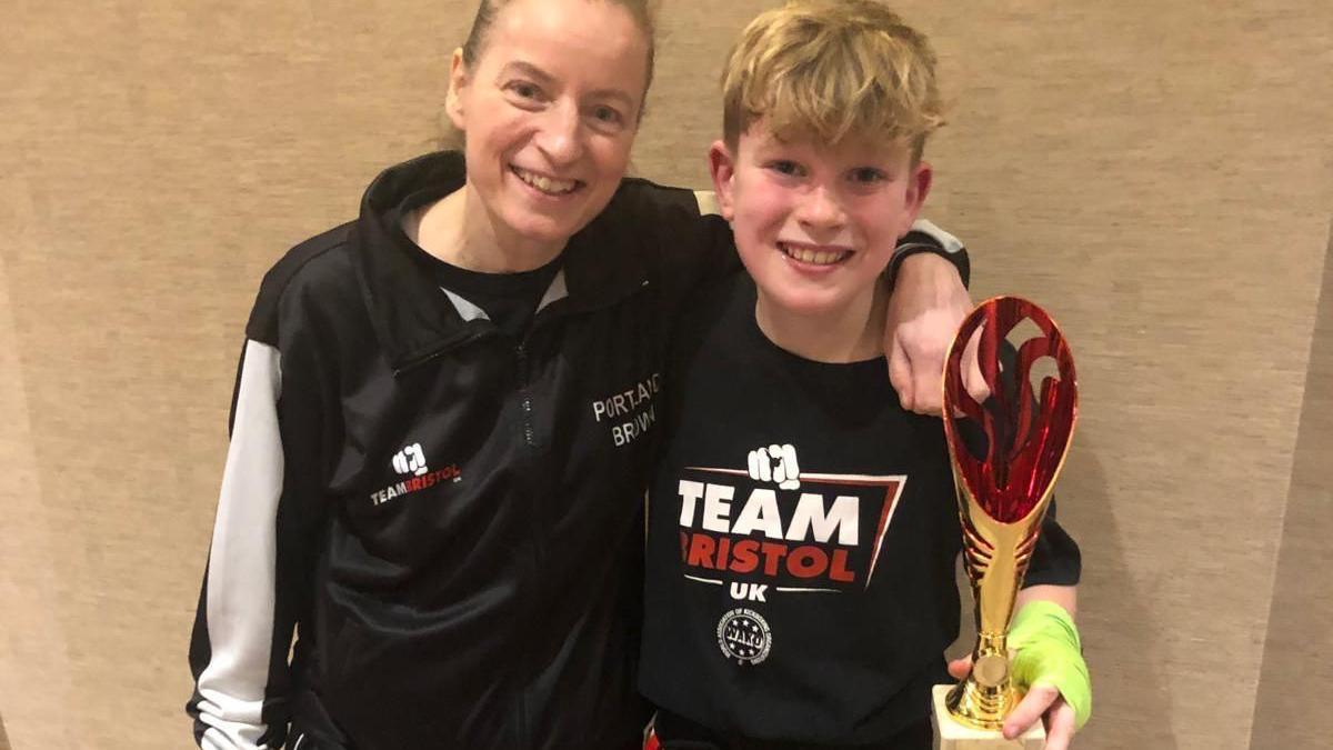 Sharon (left) with Theo (right). Both are wearing dark clothes and smiling at the camera. Theo is holding a trophy in one hand.