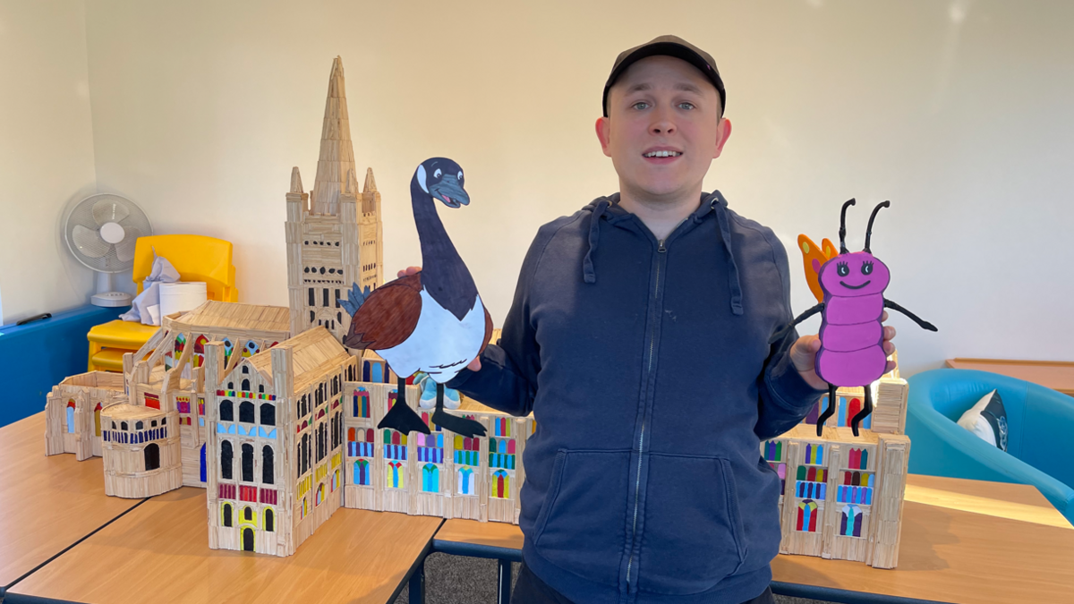 A man in a navy zip-up top and baseball cap standing in front of a model of Norwich Cathedral made out of matchsticks 