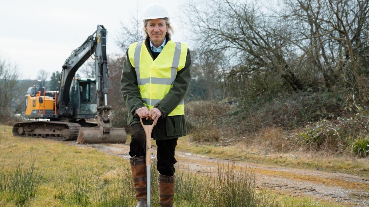 Lord Bath wearing black trousers, a grey coat, brown boots, a yellow hi-vis vest and a white hard helmet. He is holding the handle of a spade and resting his right foot on top of it, pushing it into the ground. He is smiling at the camera, and beside him is a large digger. 