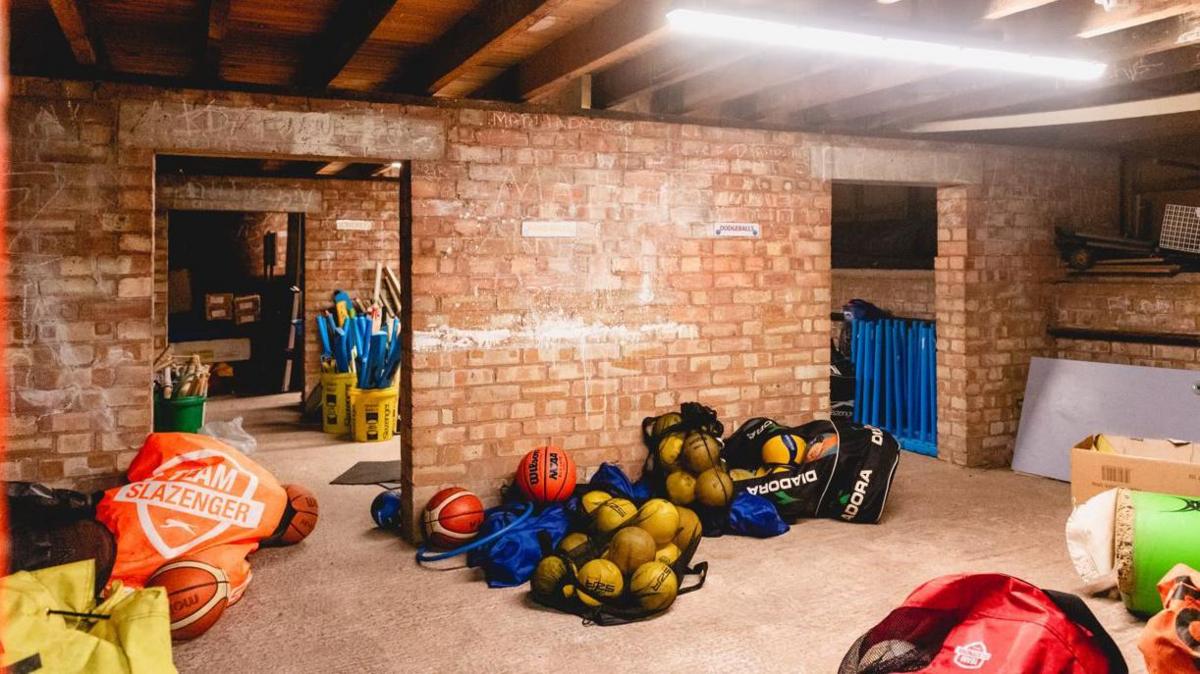 A sports storage room with brick walls and bags of balls and other sporting equipment piled up on the floor.