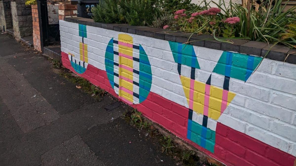 A colourful piece of wall art outside a residential property. It says JOY and has been decorated with colours of red, green, blue, yellow and pink. 