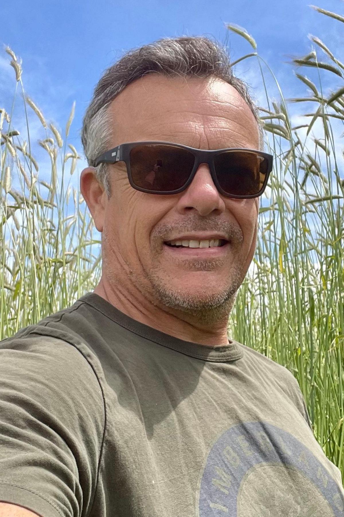 Mark Williams is standing in front of rye on a sunny day and is smiling. He is waring a pair of sunglasses and a T-shirt and has short grey hair.