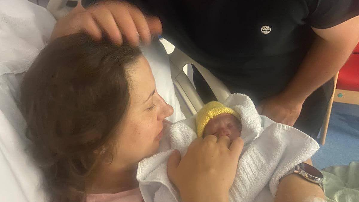 Lizzy holds her baby in her arms moments after she had been born. The baby is wrapped in white blanket and is wearing a yellow knitted had. Lizzy has lies in a hospital bed while her husband strokes her head. 