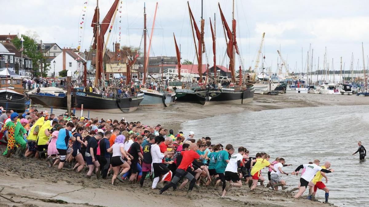 A group of about 100 people run down the mud into the River Chelmer estuary. Boats on the mudflats and buildings are in the background.