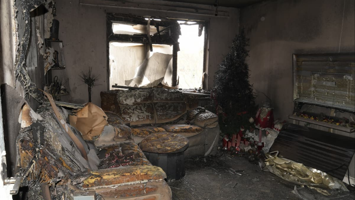 A living room, severely damaged by smoke and fire with burnt out sofas, a Christmas tree and blackened walls