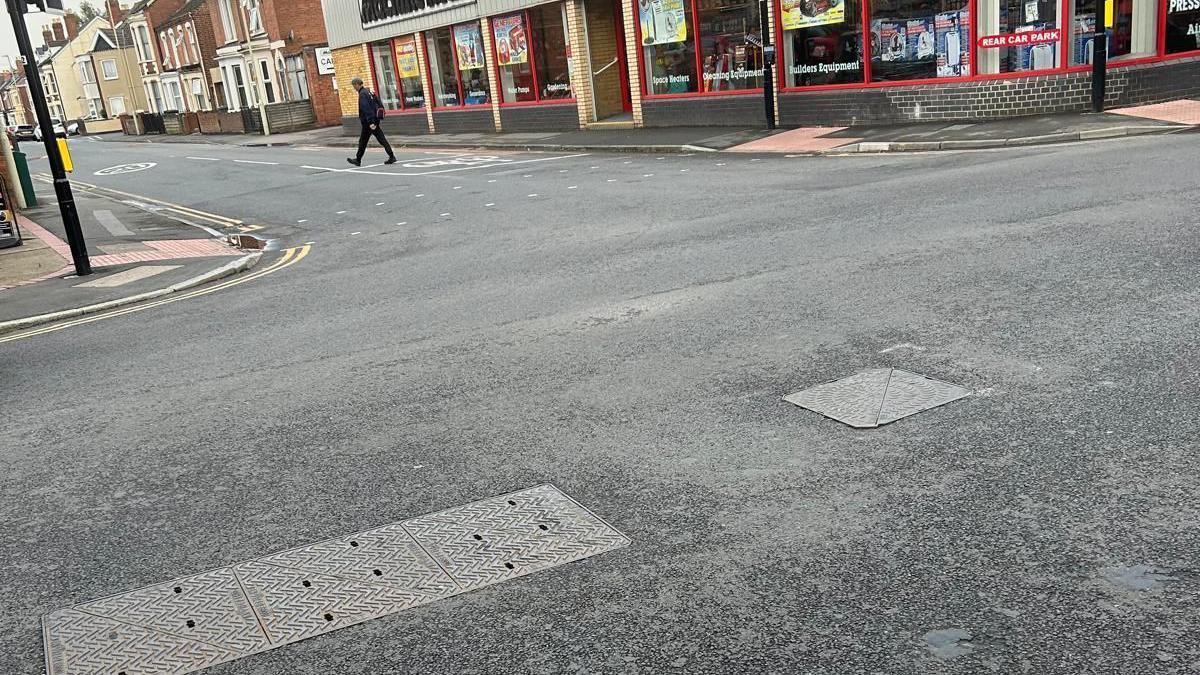 Clos-up of the manhole on a road near shops and man walking across street.