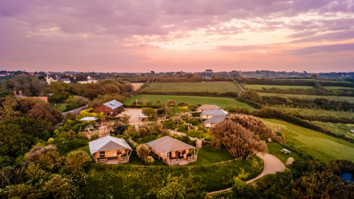 A drone shot of the campsite. The sun is setting in the background over several fields. At the front of the picture are multiple buildings which look like a mixture of tents and huts.