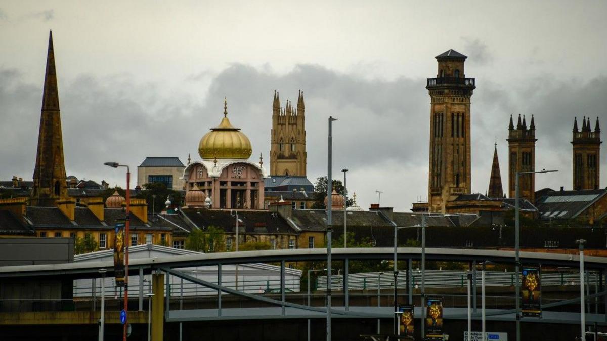 A collection of spires and the golden dome of a mosque in Glasgow. The buildings are dark stone. The sky is grey and cloudy.