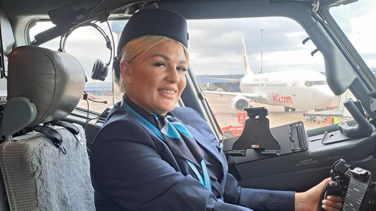 A woman in a navy flight stewardess uniform poses in the cockpit of a plane