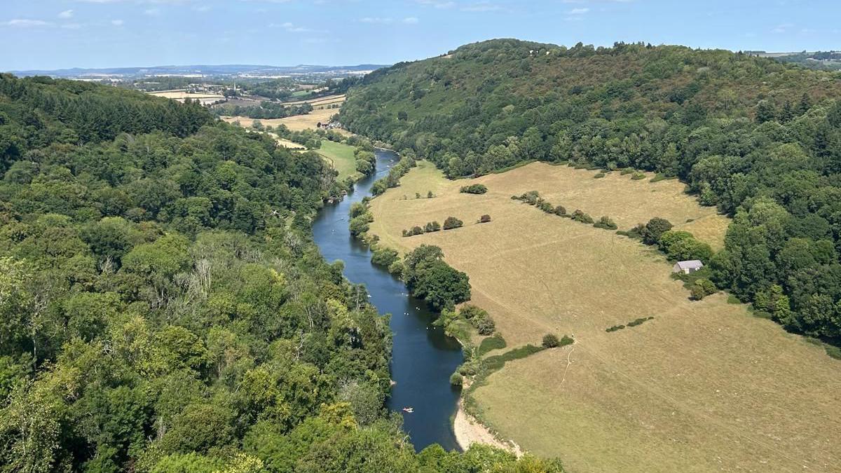 A winding river is pictured with a dense woodland to the left of it and sun scorched fields to the right of it.