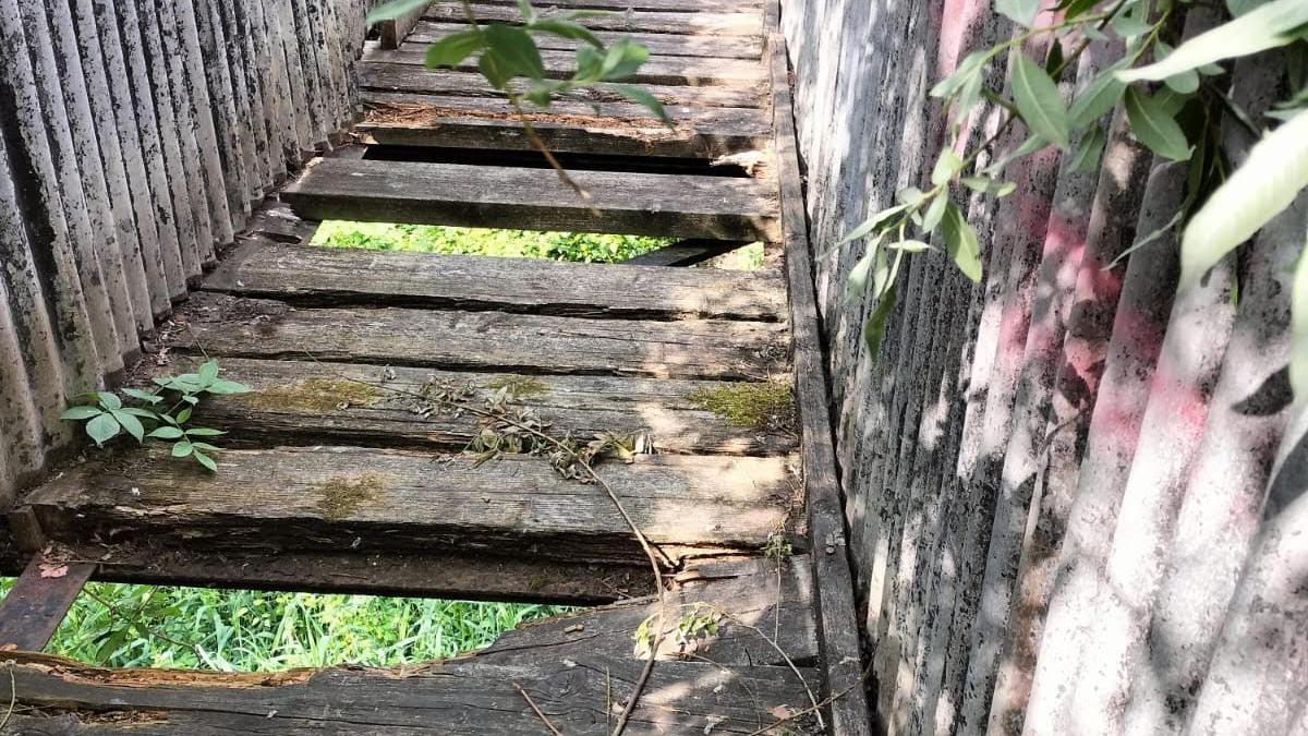 A footbridge that is falling apart. Some wooden slats are missing and you can see the ground beneath the bridge.