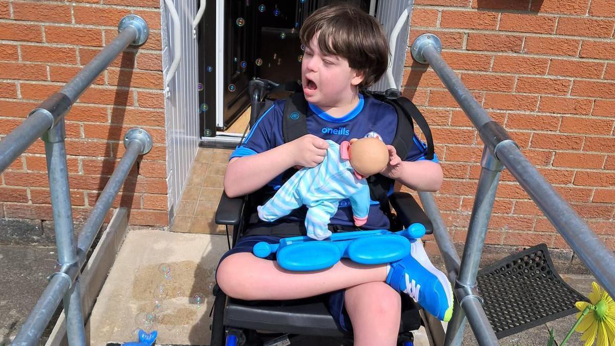 A smiling Kayden McNamee sitting in a wheelchair outside the front door of his home