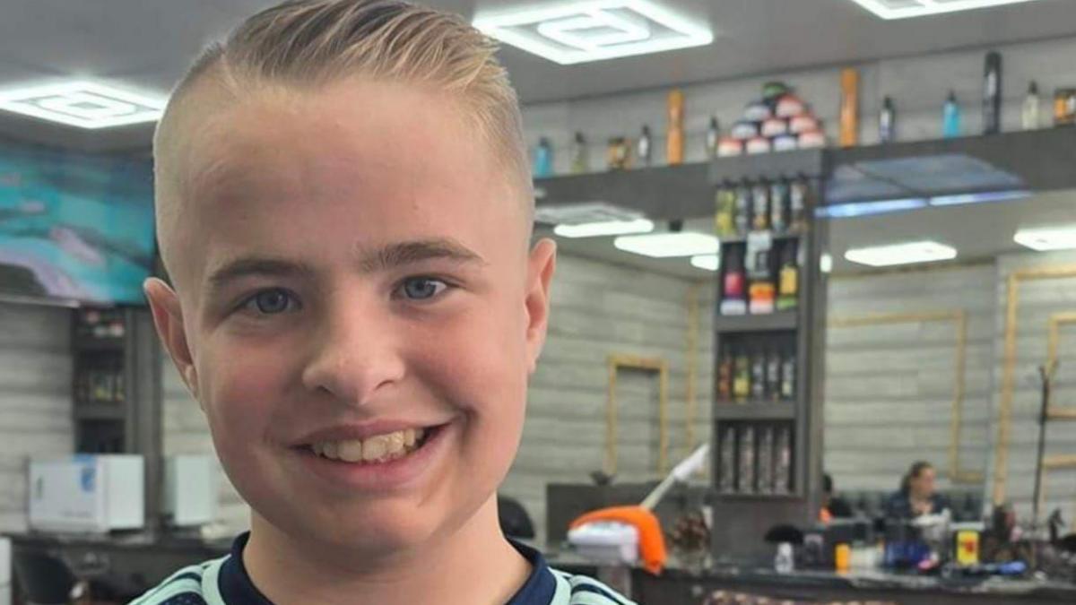 A smiling young boy with short blonde hair stands in a barbers shop.