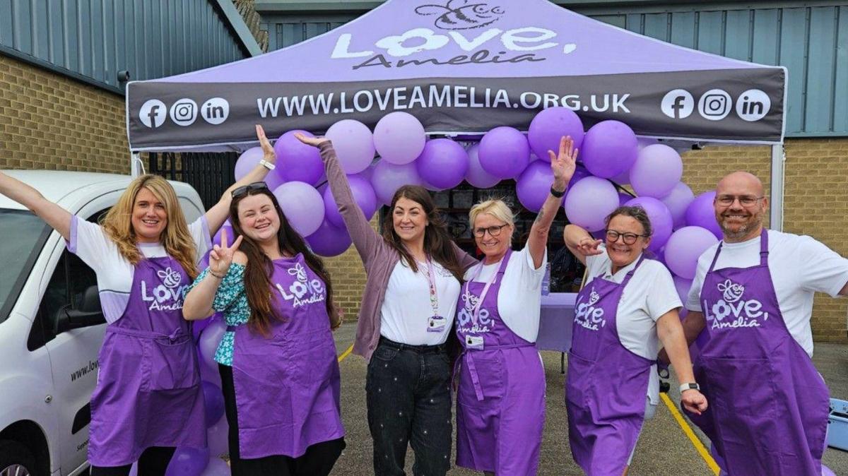 Steph Capewell, in the middle, surrounded by the Love, Amelia team. They are all wearing purple and smiling and pointing at the camera. There is an arc of purple balloons behind them. 