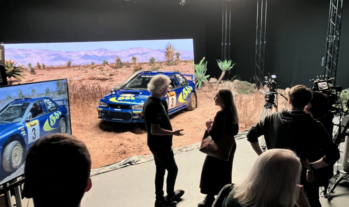 A blue rally car with 555 printed written in yellow across the bonnet, positioned indoors on a set of a sand and rocks and a bank of vegetation. At the back is a wide screen showing a similar scene of sand, dry vegetation, palms, and mountains rising in the background. In the foreground two people are talking, while others look at the set, with two cameras on tripods pointing at it.