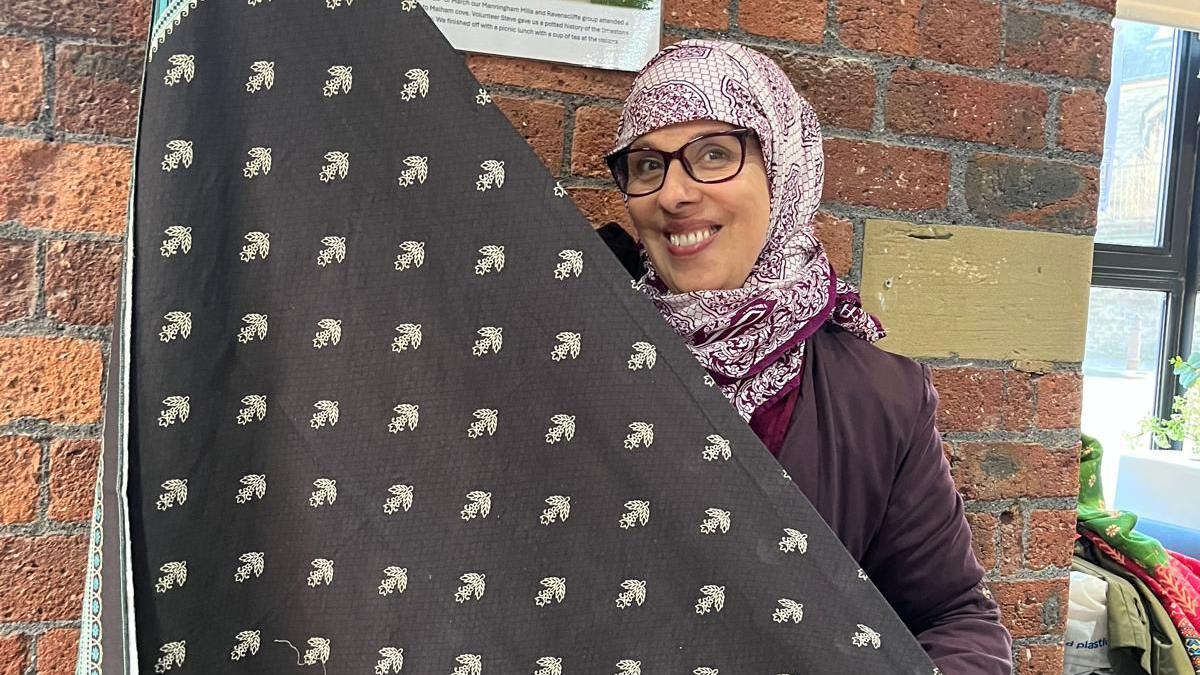 A smiling woman, wearing a headscarf, holds up a large piece of patterned black material
