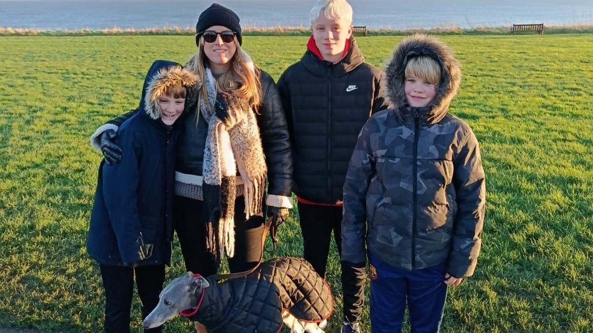 Three boys with winter coats, two are dark, one is camouflage patterned. Two have their hoods up. They are all wearing dark trousers and trainers and have short blonde hair. A lady with a hat, sunglasses and scarf also has a winter coat, dark trousers and boots. There is a lurcher dog with them with a winter coat. They are standing on a pavement with grass, water and a clear sky.