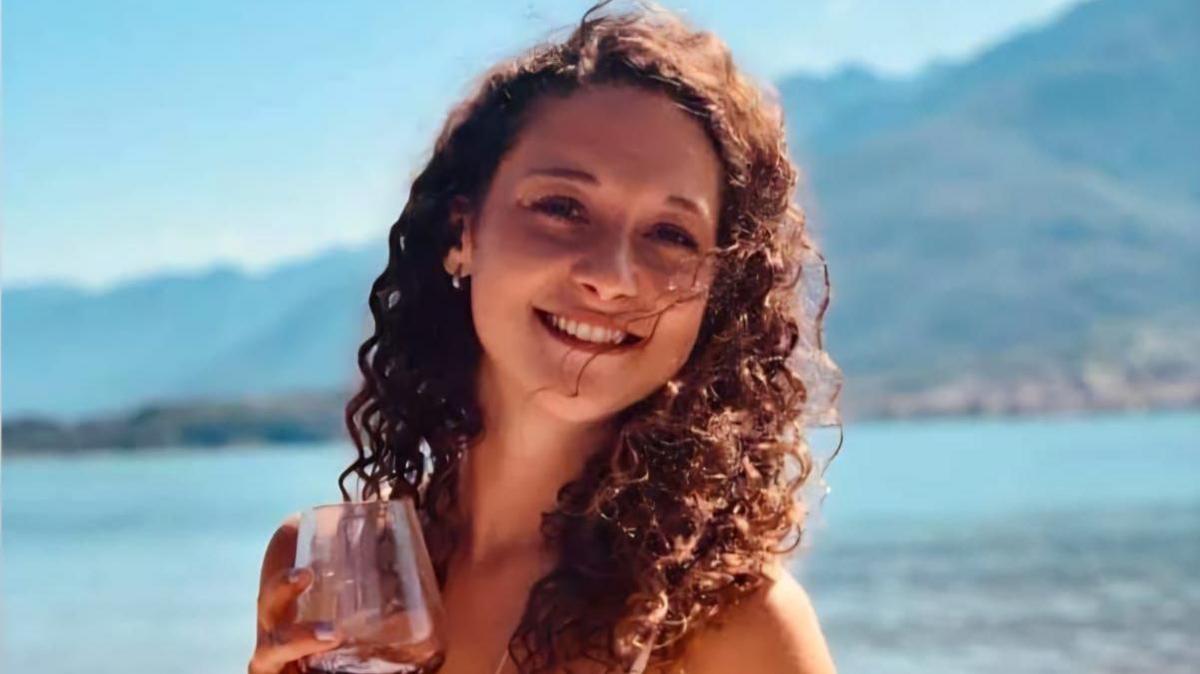 Gemma Weeks is pictured smiling at the camera and holding a wine glass on a beach. She has long, brown, curly hair and is wearing a bikini.