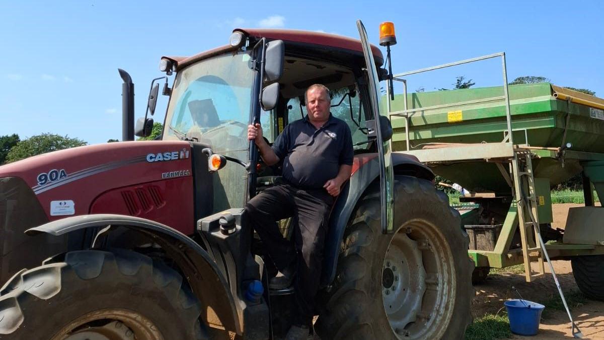 A man standing on the steps of his red tractor, looking at the camera. It has a large green implement or trailer behind it.
