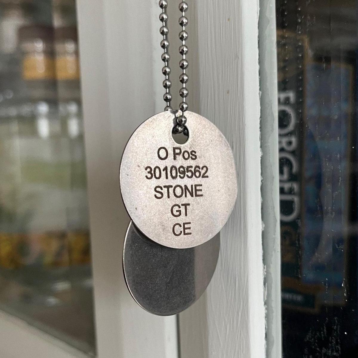 Dog tags belonging to Private Gregg Stone. Stamped on a round metal disc, attached to a chain necklace, are the words O Pos (his blood group), 30109562 (his Army number), Stone GT, CE (for Church of England).