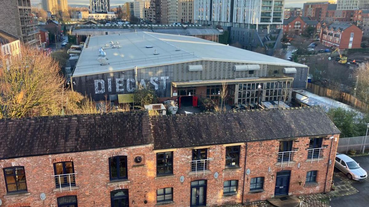 The Diecast complex as seen behind a set of redbrick terraced buildings, with the skyscrapers of Manchester's skyline seen in the distance.