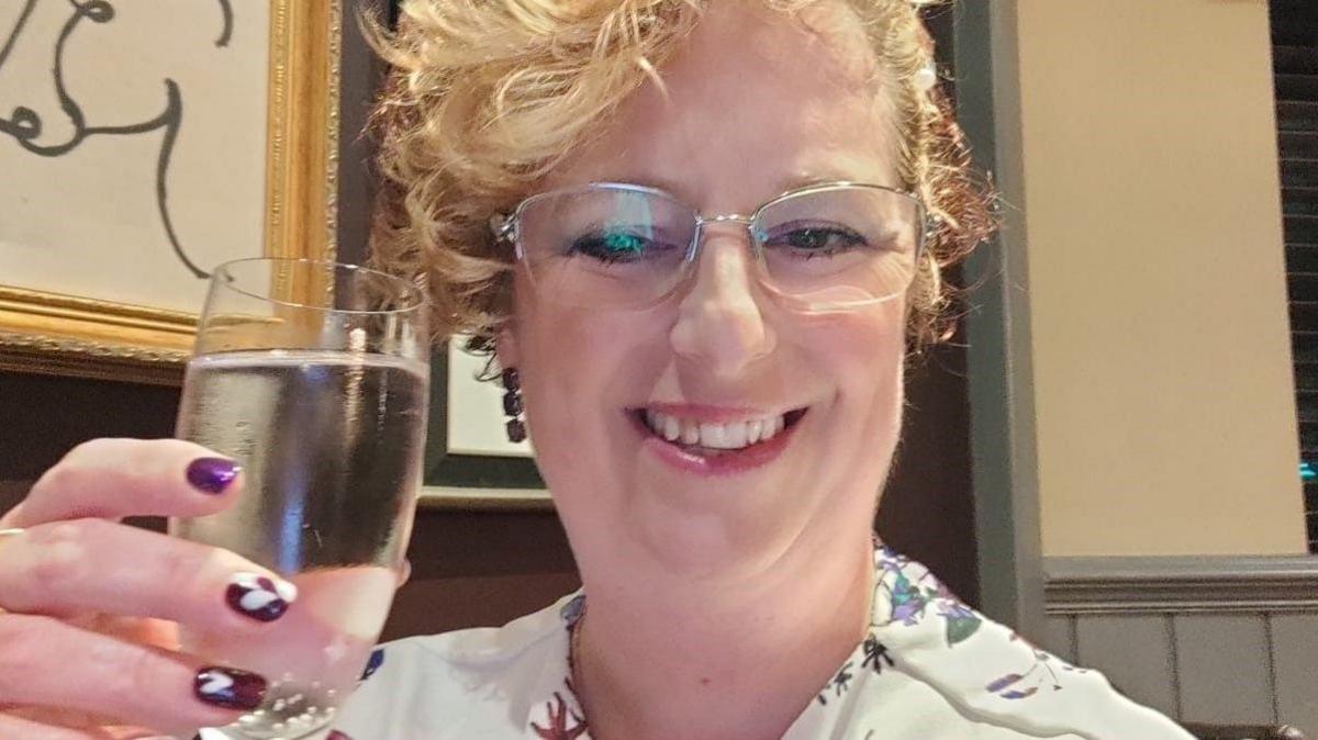 Sarah Wilkinson with her fair hair swept up and curling over her right forehead, wearing glasses, smiling at the camera and toasting herself