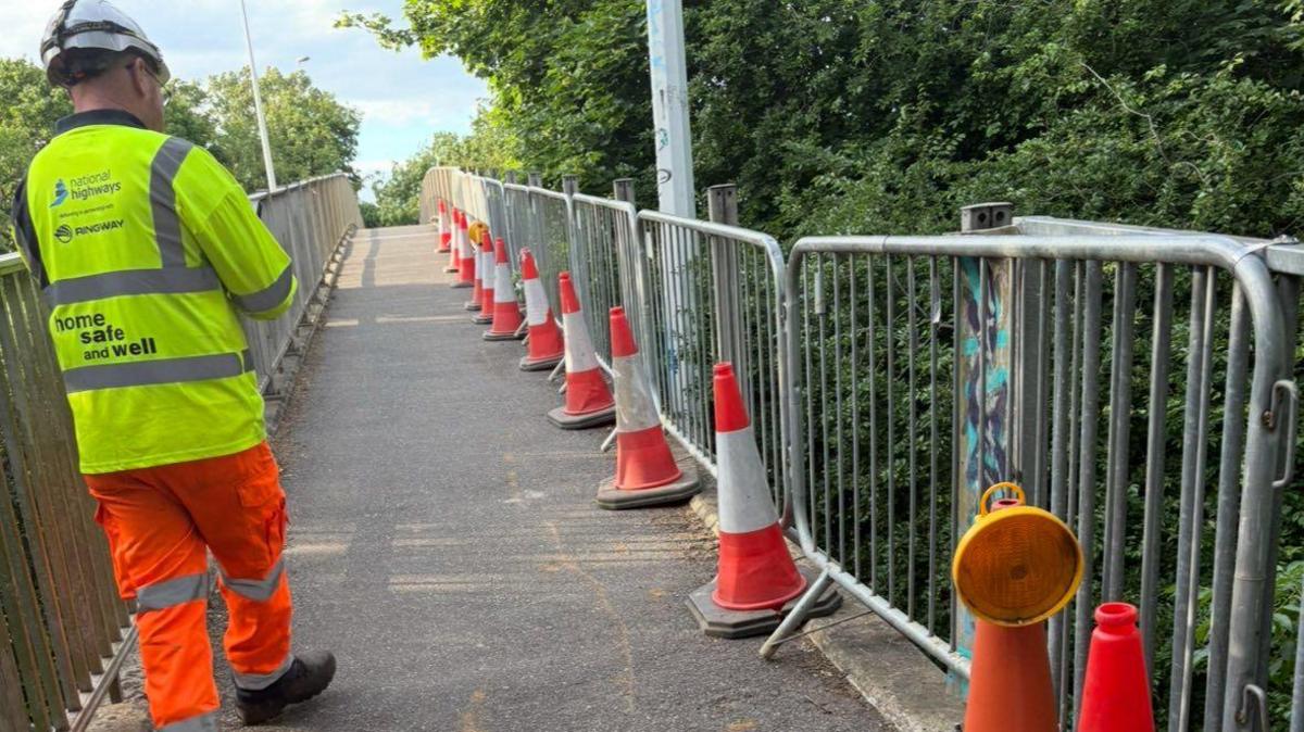 A man from National Highways, wearing a branded hi-vis jacket and trousers, walking along the bridge with his back to the camera.
