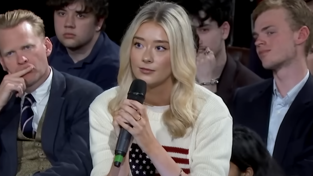 Tilly Middlehurst a young woman standing at a wooden table and holding a microphone. She has blonde hair and is wearing a cream jumper with the flag of the United States on it.