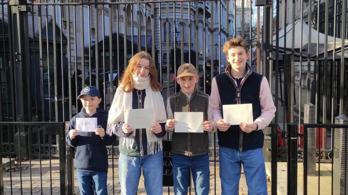 Four children stand outside the black gates of downing street. The three boys and one girl and holding up their letters on A4 paper in front of them. 