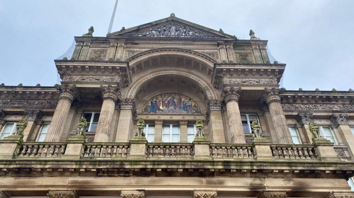 An external shot of the upper floor Birmingham Council House, which is a sandstone building with moulded columns and a balcony. Statues of lions line the balcony on plinths and a central arch sits behind it, featuring a mosaic. A pale blue sky can be seen above.