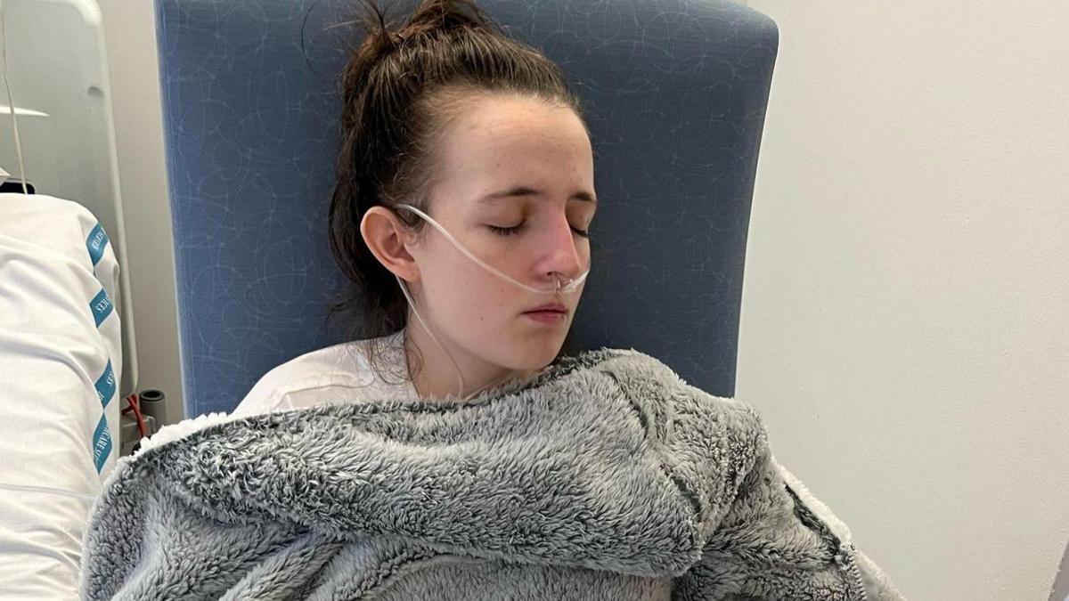 Amy Symington is sitting in a high-backed chair in hospital. She has an oxygen tube attached to her nose. She has a thick, grey fleece blanket wrapped around her and is dozing. To the left we can see the side of a hospital bed.