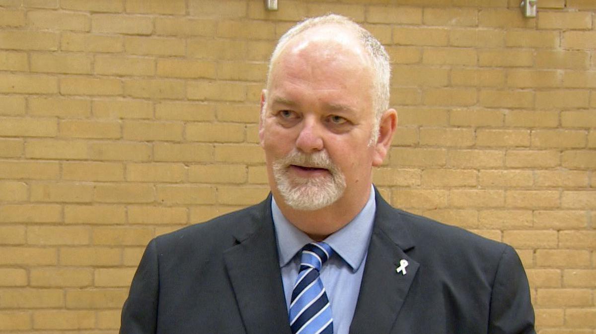 The Humberside Police and Crime Commissioner, Jonathan Evison, stands in front of a cream brick wall. He is wearing a dark suit with a blue shirt and blue striped tie. Mr Evison has balding grey hair and a grey goatee beard.