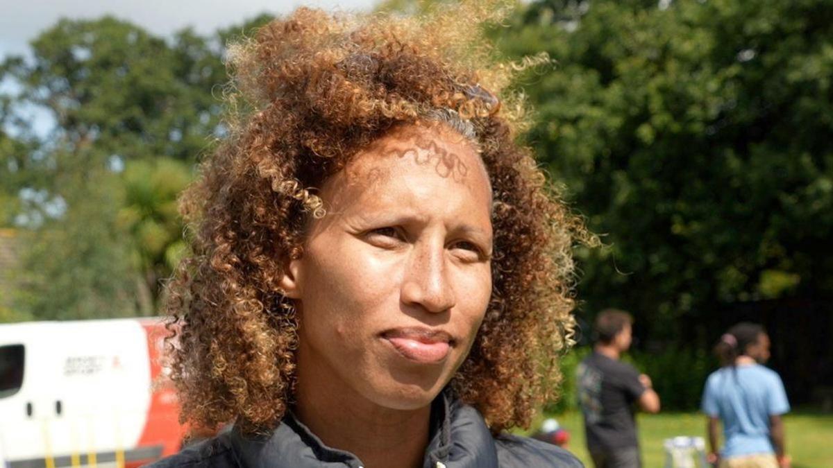 Serena looks at the camera as people set up the sports day behind her. She is wearing a white jumper with blue stripe hoops with a black gillet. Her hair is slightly blowing in the wind.