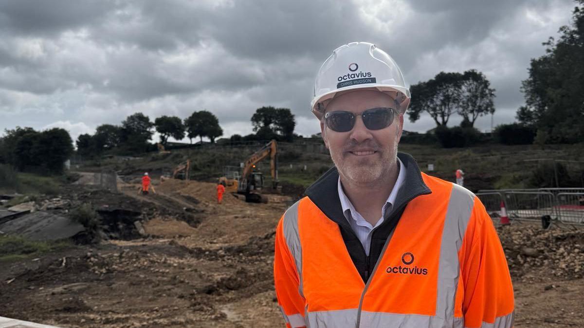 Chris Hudson in high-vis clothing, hard hat and sunglasses looking into camera with a building site behind