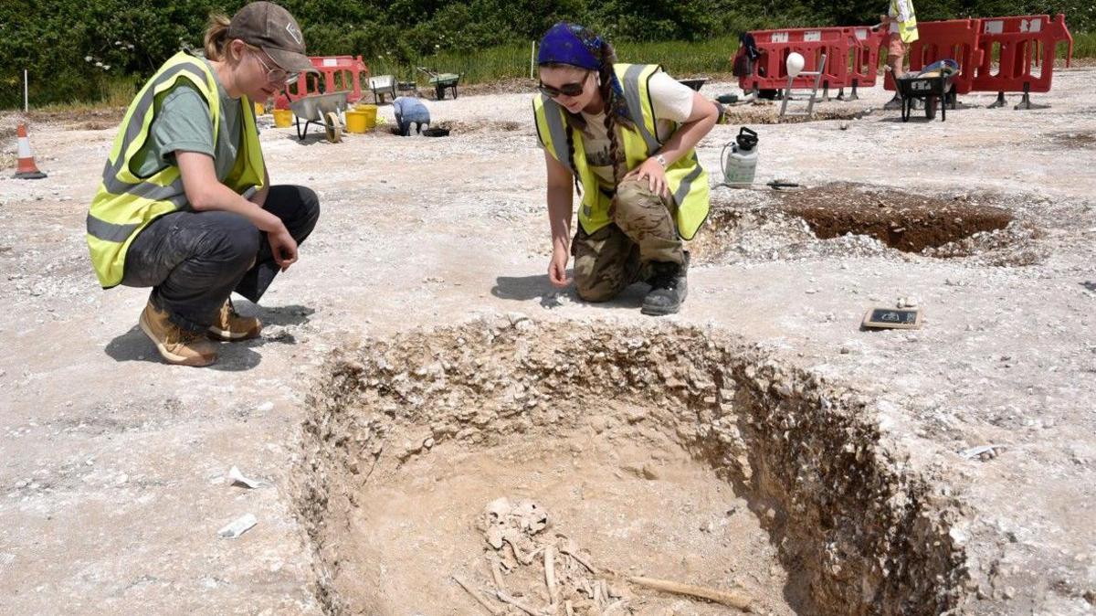 Two women wearing hi-vis vests and peaked caps crouching over an excavation pit containing a skeleton