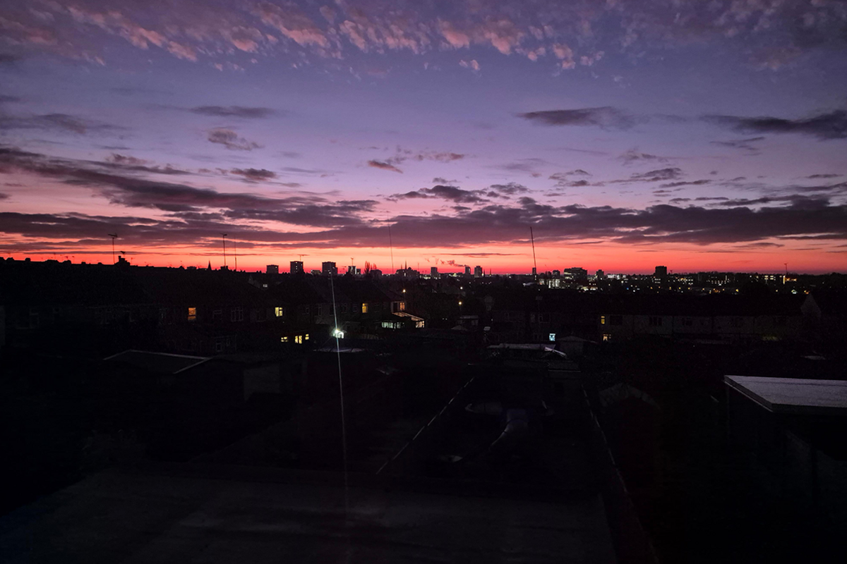 An evening sky which runs from dark blue through darker clouds to red and yellow at the horizon as the sun sets. Underneath, the lights from dark houses dot the landscape.