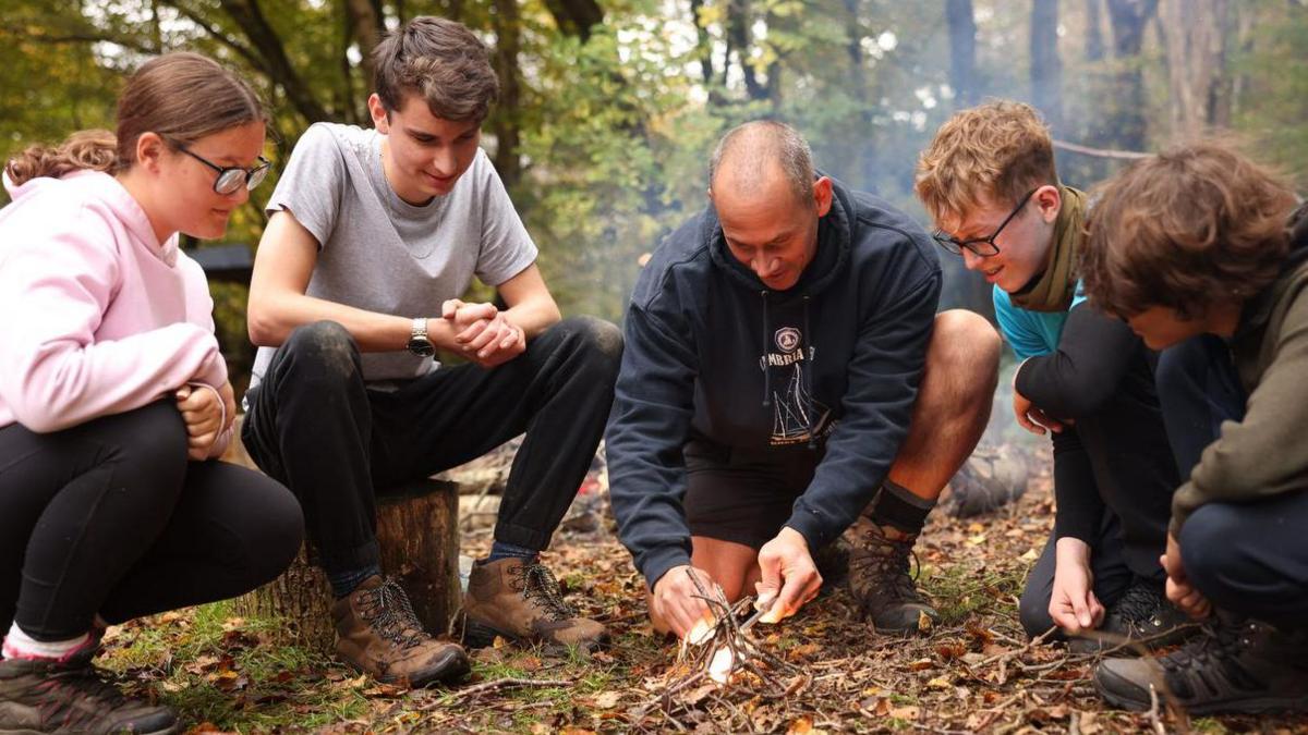 A man lights a fire using some twigs in woodland as four teens crowd around him. 