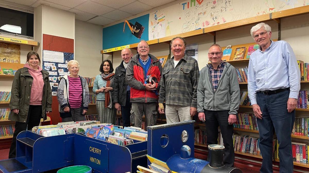 The society trustees are standing in a line inside the library. There are full bookshelves behind them and they are all smiling. There are three women, to the left, and five men, stood to the right.