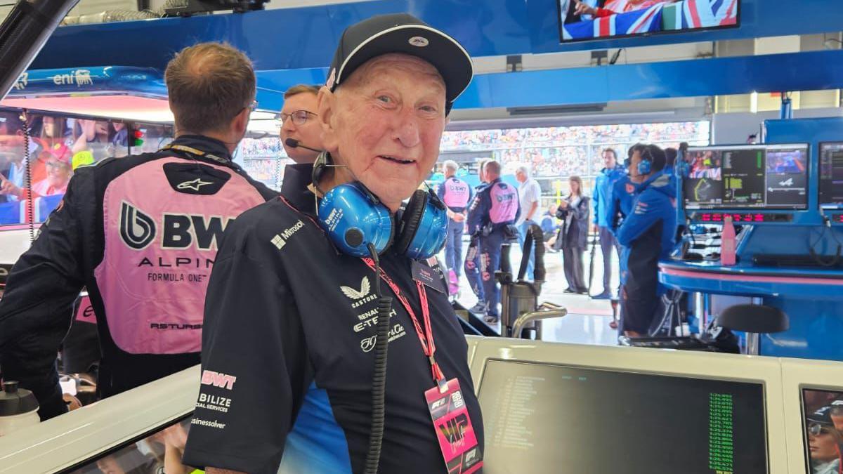 An older man wearing a black cap, blue headphones, and a black shirt stands in a motorsport pit area. Behind him, team members in pink and black uniforms work near monitors, with spectators visible through the open garage.