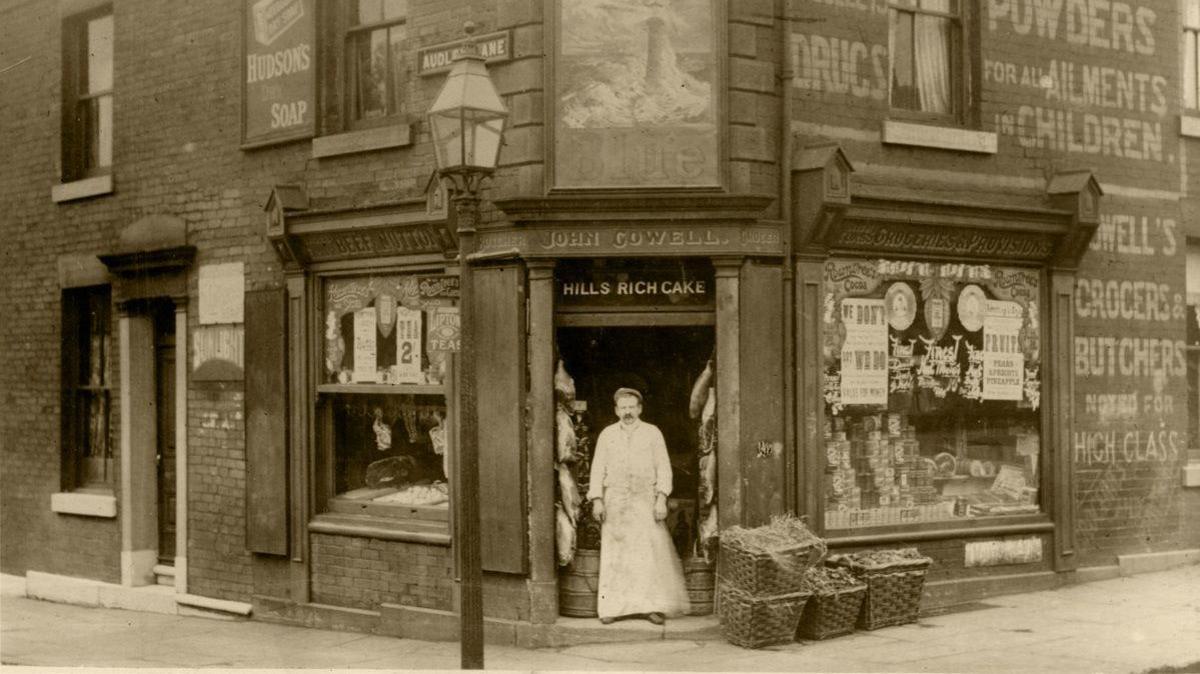 An old photograph from the 1800s of the Hills cake shop. A man in a white frock with a moustache can be seen stood in the doorway of the shop, with wooden baskets placed in front.